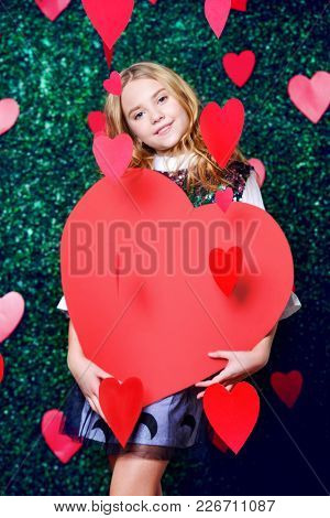 Happy pre-teen girl holding red heart surrounded by little hearts over lawny background. First love. Valentine's Day. 