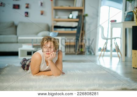 White Big Room With A Wooden Shelf On The Background. A Boy Is Lying On A Capret In The Middele Of T