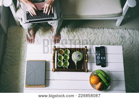 Senior Man Typing On Laptop With His Feet On Fluffy Rug, Table With Tea Set, Table Computer, Fruits 