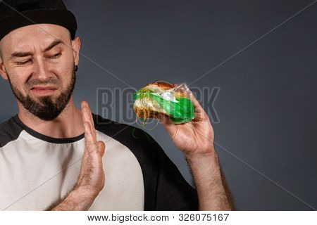 The Concept Of Fast Food And Ecology. A Man In A Cap And A Beard, Holding A Hamburger With A Plastic