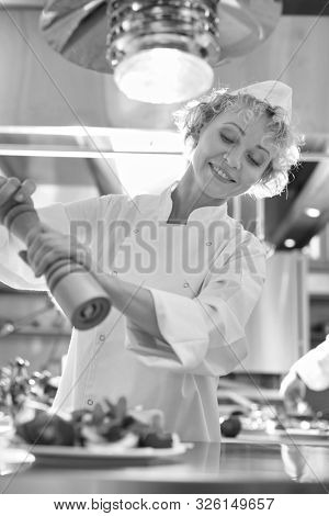 a black and white photograph of a Mature chef using peppermill on salad in plate at restaurant