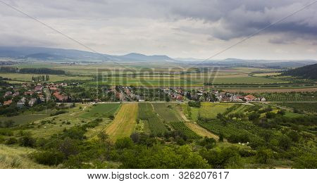 Small Village In A Valley. Hills, Mountains And Fields Around. Dark Cloudy Summer Day. Picture Taken