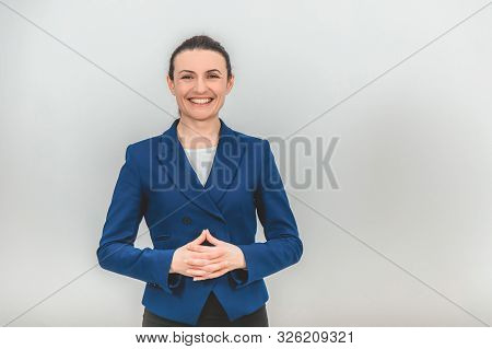 Female Young Teacher Standing, With Her Fingers Interlaced, Looking At The Camera, Smiling.