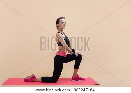 A Young Attractive Girl With A Ponytail, A Brunette, Stands On One Knee, Doing Stretching On A Pink 