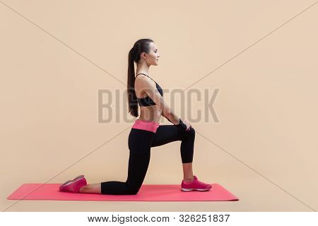 A Young Attractive Girl With A Ponytail, A Brunette, Stands On One Knee, Doing Stretching On A Pink 