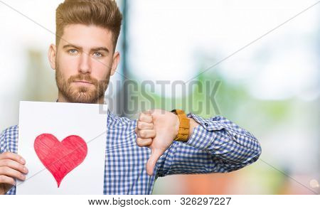 Young handsome man holding paper with red heart with angry face, negative sign showing dislike with thumbs down, rejection concept