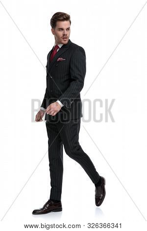 side view of elegant businessman wearing double breasted suit and red tie, looking to side, walking isolated on white background in studio