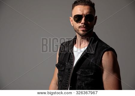 Eager man looking forward while wearing sunglasses and a black jeans vest, standing on gray studio background