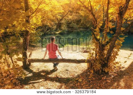 Man sits on the branch of tree near river at auntumn forest. Nature landscape scene.
