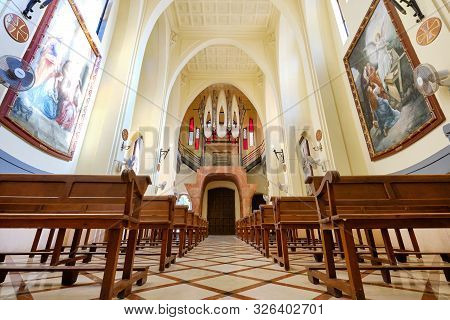 Novelda, Spain- October 3, 2019: Aisle Between Wooden Pews Leading To Pipe Organ Musical Instrument 