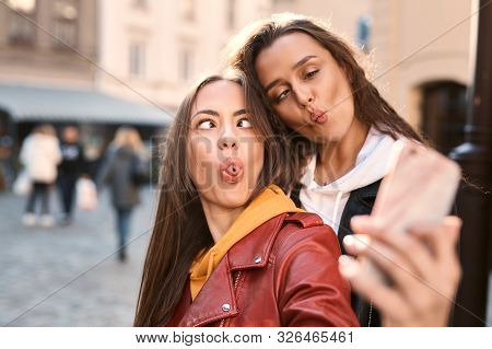Portrait Of Cute Young Women Making Funny Faces For Picture On Phone Uotdoors In City.