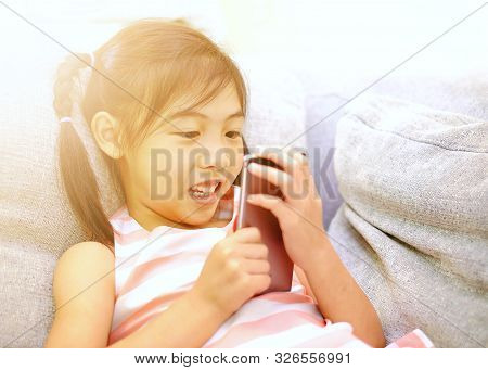 A Young Asian Girl Playing With Her Smartphone On A Sofa. Sun Flare.