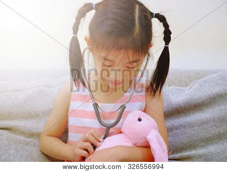 An Asian Girl Playing Doctor With A Stethoscope On Her Pink Rabbit, Smiling.