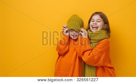 Winter portrait of happy children wearing knitted hat, snood and sweaters. Girls having fun, playing and laughing on yellow background. Fashion concept.