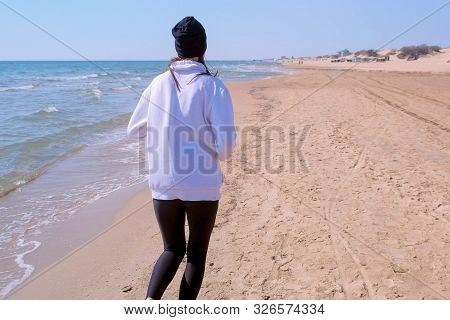 Barefooted Woman Is Gogging On Sea Sand Beach, Back Vew. Girl Is Wearing Leggings, Hoodie And Hat Ru