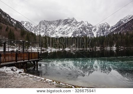Lake With Green Forest And Snowy Mountains In The Background And Skyfall Effect. Italian Lake During