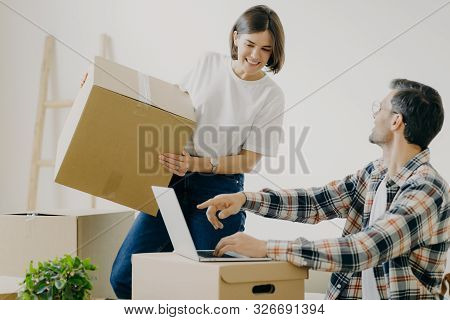 Happy Young Man Points Into Laptop Computer, Searches New Design For Flat, Busy Woman Carries Boxes,
