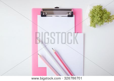 Three Pencils Lie On A Pink Clipboard And A Sheet Of Paper. In The Frame You Can See A Plant In A Ca