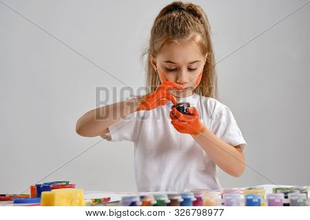 Little Girl In White T-shirt Sitting At Table With Whatman And Colorful Paints On It, Painting Her H