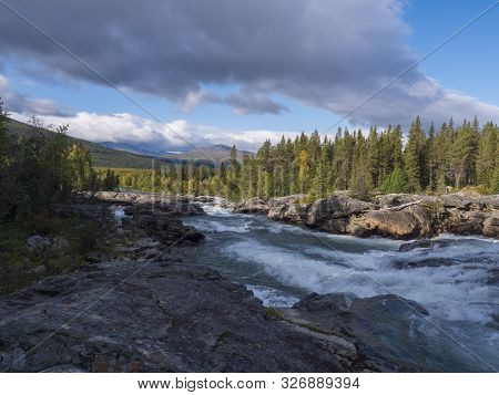 Beautiful Northern Landscape With Wild Glacier River Kamajokk, Boulders And Spruce Tree Forest In Kv