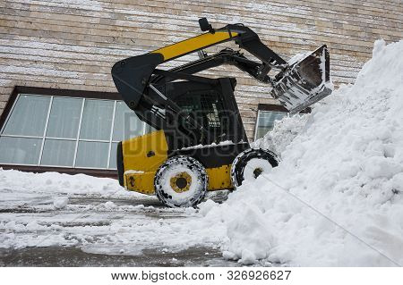 Minitractor Shovels Snow In A Big Pile. The Background Of The Image Is The Wall Of The Building. Sel