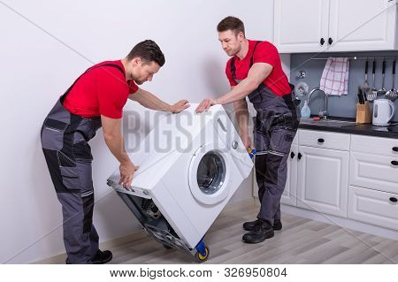 Two Young Male Professional Movers In Uniform Placing The Modern Washing Machine In The Kitchen