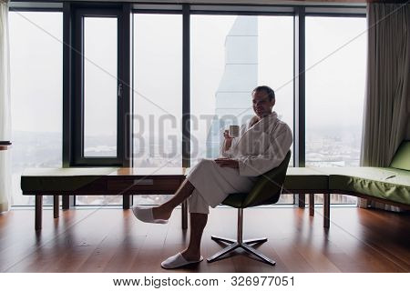 Young Handsome Carefree Man Wearing Bathrobe Near Modern Full Length Window Enjoying A Cup Of Coffee
