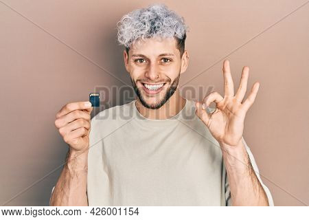 Young hispanic man with modern dyed hair holding sdxc card doing ok sign with fingers, smiling friendly gesturing excellent symbol 