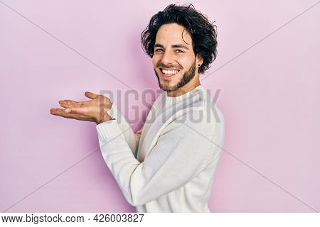 Handsome hispanic man wearing casual white sweater pointing aside with hands open palms showing copy space, presenting advertisement smiling excited happy 