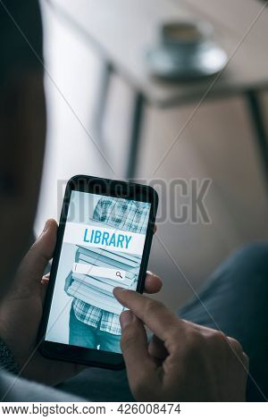 a young man, sitting at a coffee table in his living room, is searching an ebook to lend on an online library with his smartphone, with a simulated search engine in its screen