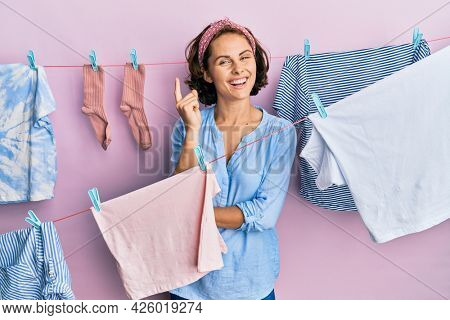 Young brunette woman doing laundry around string hangs with a big smile on face, pointing with hand and finger to the side looking at the camera. 