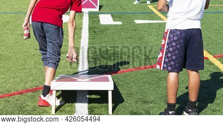 Rear View Of Two Boys Playing Cornhole Bean Bag Toss Game On A Turf Field.