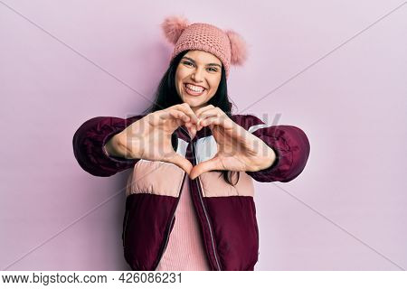 Young hispanic woman wearing wool winter sweater and cap smiling in love doing heart symbol shape with hands. romantic concept. 