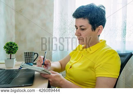 Woman With Short Hair And Gray Hair Makes Notes In Paper Notebook While Sitting At Table In Front Of