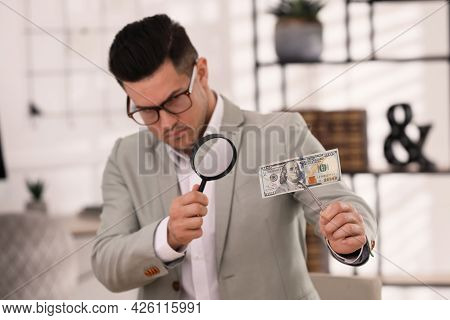 Expert Authenticating 100 Dollar Banknote With Magnifying Glass In Office, Focus On Hand. Fake Money