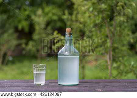 Big Bottle And Glass Of Moonshine Or Vodka On Wooden Table In Sunny Summer Day In Garden, Close Up