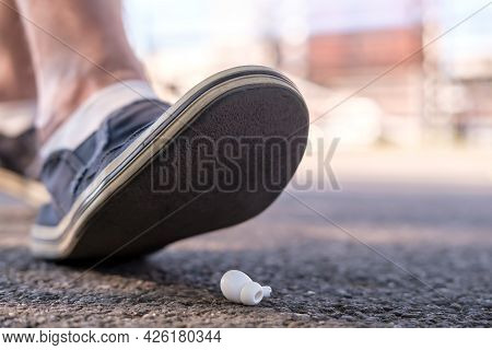 Foot Of A Man In Shoes Steps On A Lost Wireless Headphones, Which Lies On The Asphalt Sidewalk, Outd