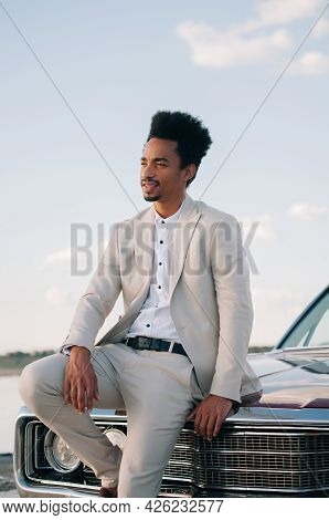 The Groom Of A Young African American At Sunset In Nature Leaned His Hands On A Red Convertible.