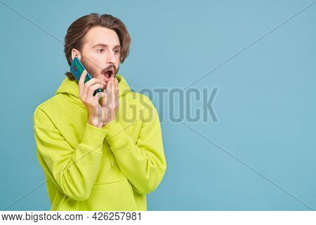 Funny young man in light green hoddie is talking on the phone and is very surprising. Studio portrait on a blue background with copy space.