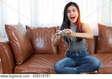 Vdo Game Console Station Concept. Active Asian Woman Sitting On Sofa, Holding Joystick And Playing E