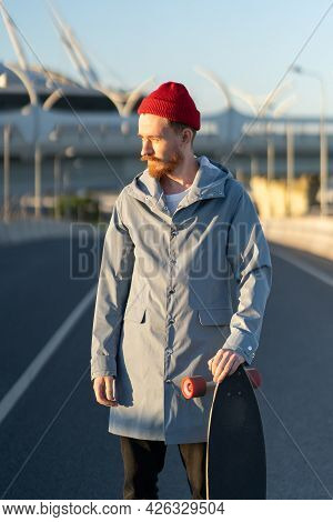 Casual Man With Longboard On Sunset Over Modern City Building. Hipster Skater Holding Skateboard Wea