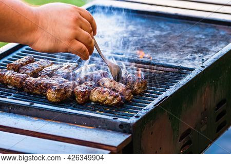 Preparing Meat Rolls Called Mici Or Mititei On Barbecue. Close Up Of Grill With Burning Fire With Fl