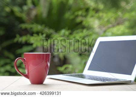 Good Morning With Coffee In Red Cup With Computer Laptop, Green Garden Bokeh Background. Copy Space.