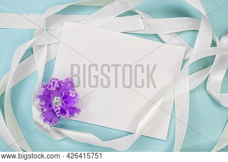 On A Blue Background Among A White Ribbon There Is A Clean Paper Sheet With A Violet Flower. A Layou