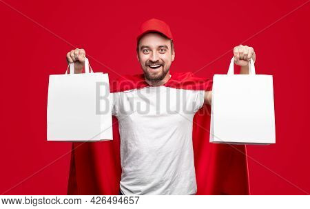Cheerful Bearded Delivery Man In Red Cap And Superhero Cloak Demonstrating Blank White Paper Bags Wh