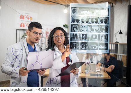 Group Of Two Multiracial Doctors, Standing Behind Glass Wall, Using Laptop While Examining Tomograph