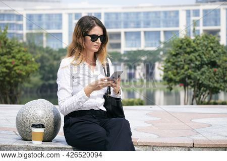 Serious Frowning Businesswoman Sitting Outdoors With Cup Of Coffee And Aswering Text Messages On Sma