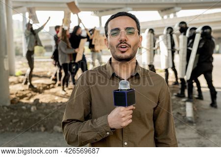 Portrait of young mixed race journalist with beard speaking into microphone while covering riot: protestors arguing with riot police in background