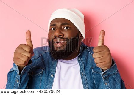 Close-up Of Awkward Black Man In Beanie Showing Thumbs-up But Feeling Cringe, Standing Unsure And Wo