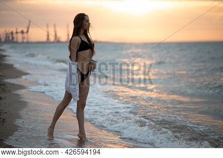 Young Girl In A Whight Shirt Walks On The Sea Coast At Sunrise
